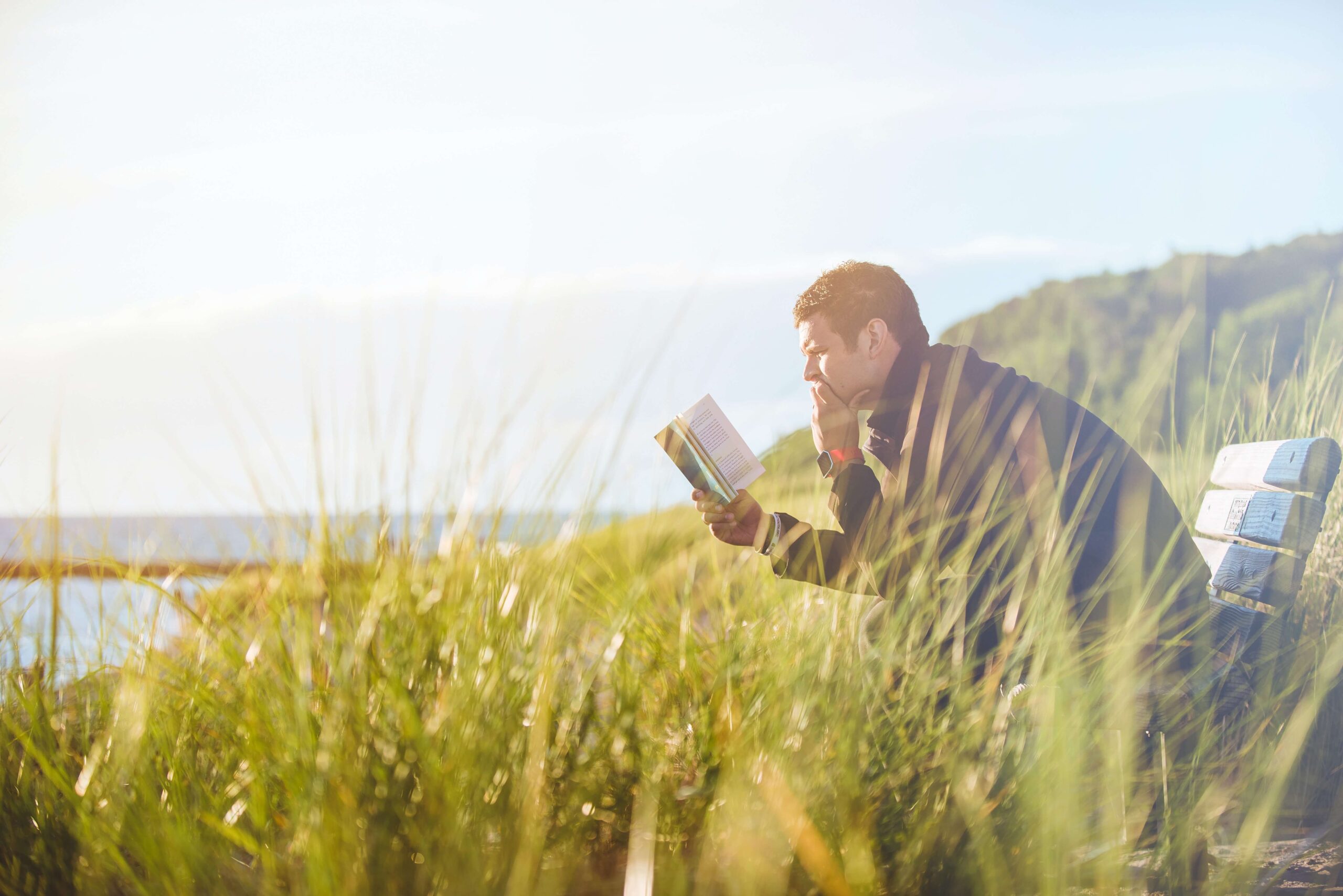 A man sitting on a bench in tall grass, facing the water, reading a book